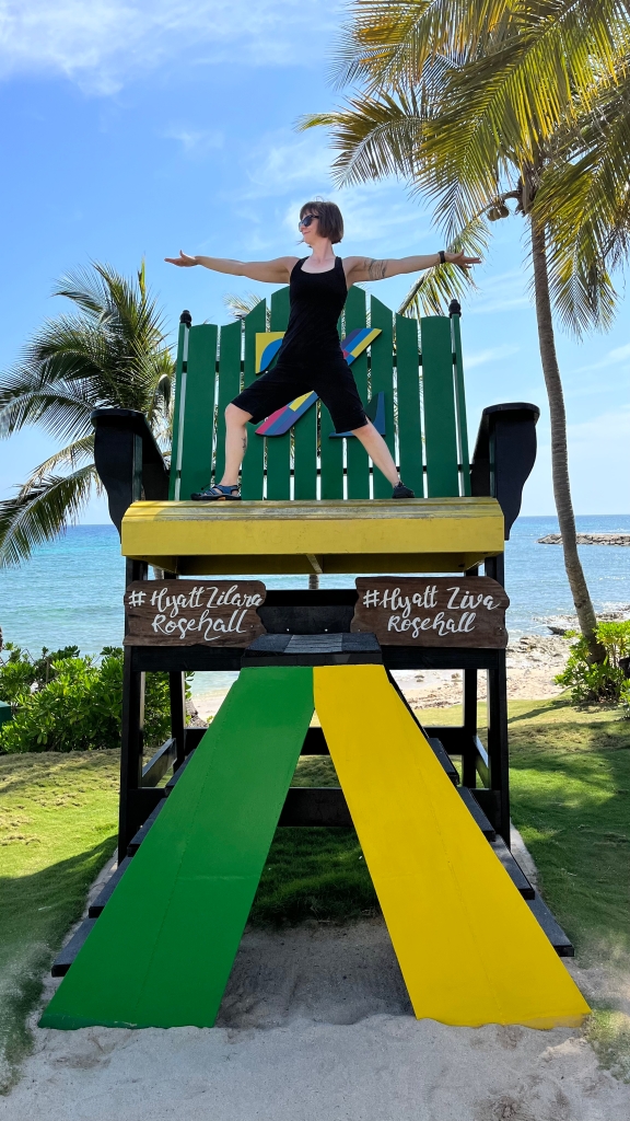 Kate in Warrior 2 pose in front of a Jamaican beach. She's standing on a large, decorative chair meant for such photos. 